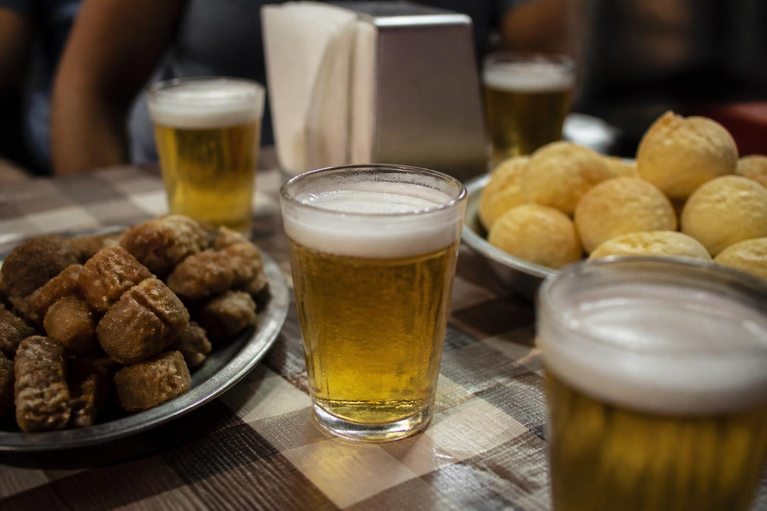 Mesa de boteco com copos de chope gelado, torresmo e pão de queijo